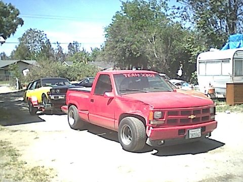 1987 Ford Bronco - A red pickup truck tows a yellow car on a trailer, with a trailer home and trees in the background.