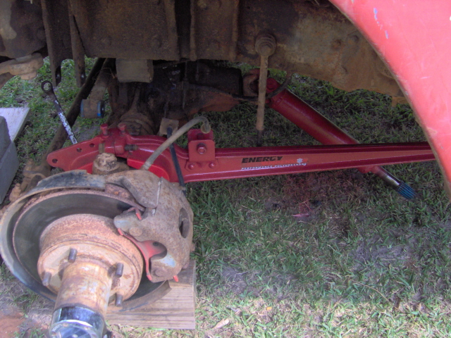 Red suspension arm with "ENERGY" logo and "Performance Suspension Components" text, attached to a rusty brake assembly