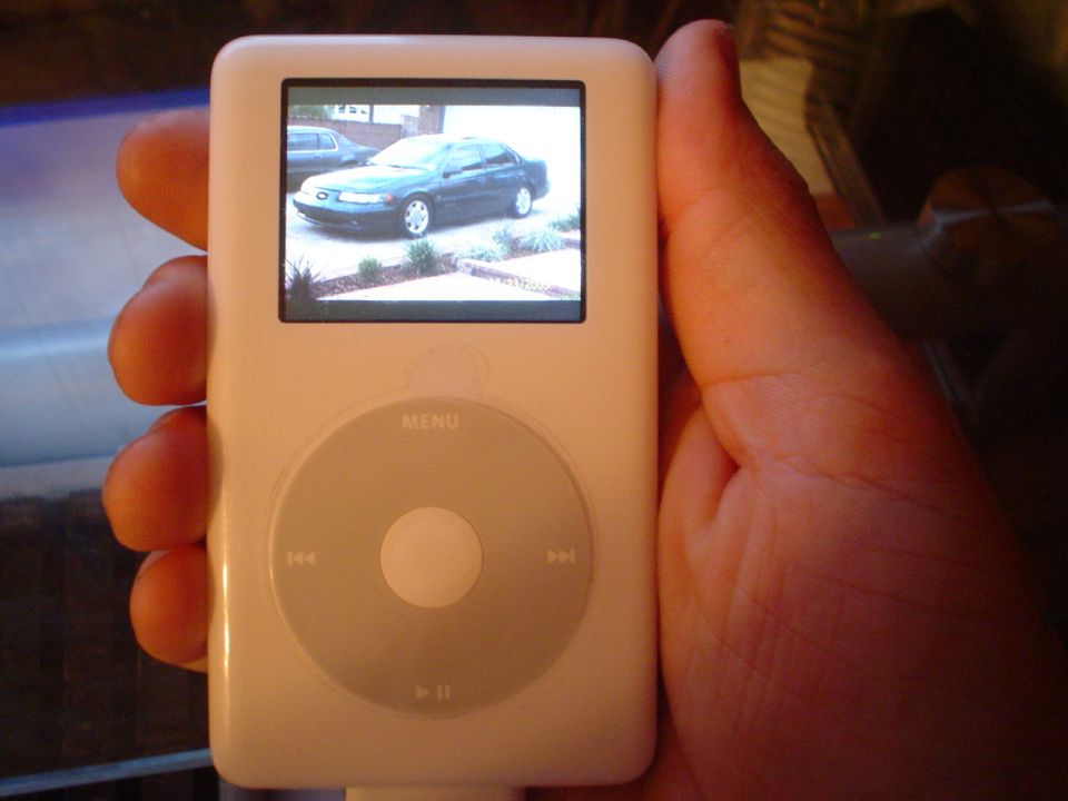 Hand holding a white iPod Classic displaying a photo of a dark car on its screen
