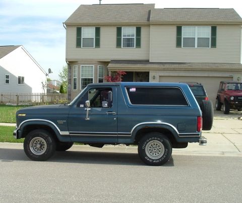 1982 Ford Bronco - A side view of a dark blue Ford Bronco is parked on a residential street in front of a house.