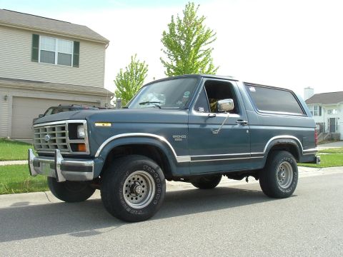 1982 Ford Bronco - This blue Ford Bronco is parked on a suburban street with a house and trees in the background.