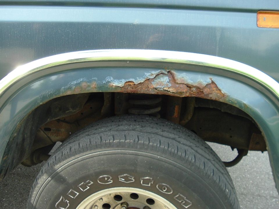 Rusted-through rear wheel arch lip on a dark green Bronco with Firestone tire visible