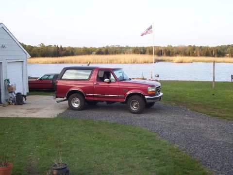 1995 Ford Bronco - A red 1995 Ford Bronco is parked on a gravel driveway next to a house, with a body of water and trees in the background.