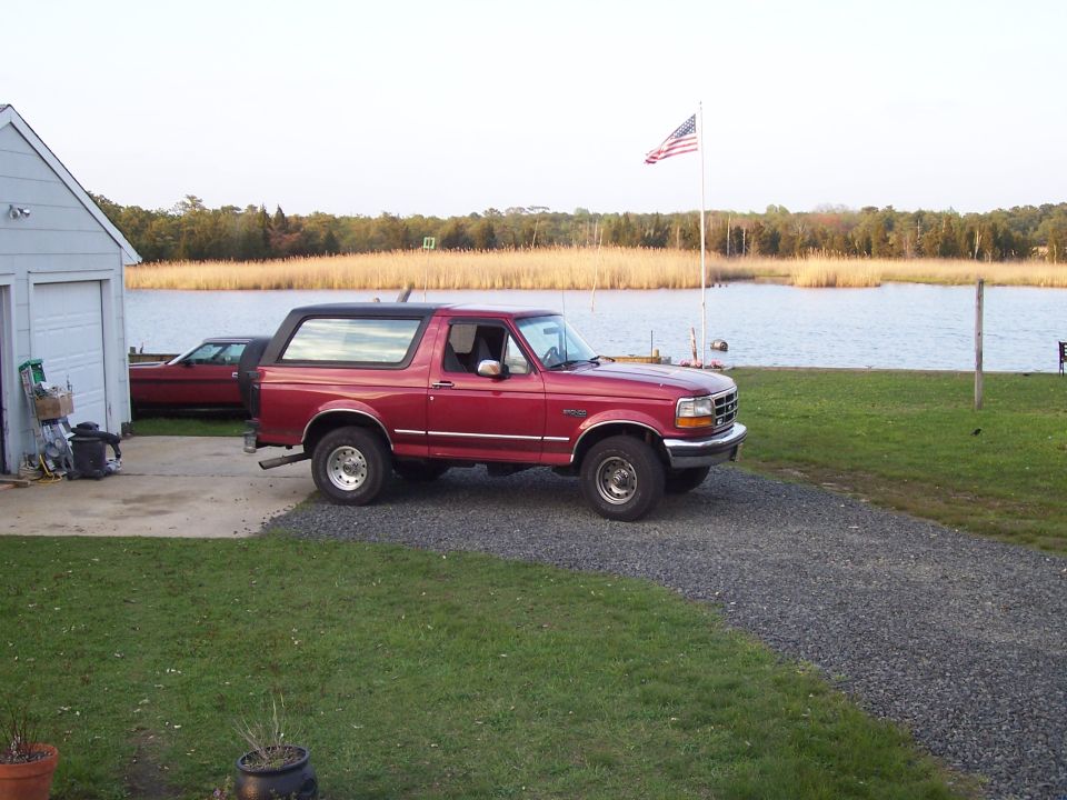 Red 1995 Ford Bronco parked on gravel near a lake, with an American flag flying