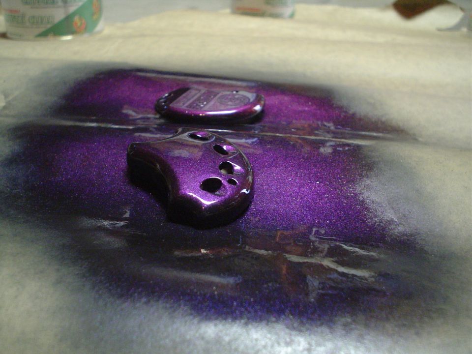 Two purple key fobs with holes, one with a metallic finish, on a surface with overspray
