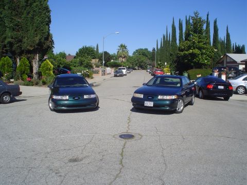 1995 Ford Taurus - Two dark green Ford Taurus sedans are parked on a sunny residential street.