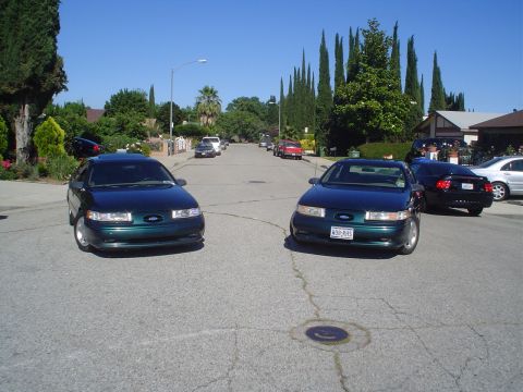 1995 Ford Taurus - Two dark green 1995 Ford Tauruses are parked on a suburban street under a clear blue sky.