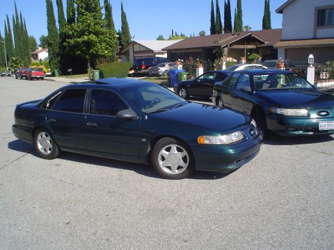 1995 Ford Taurus - Two dark green sedans are parked on a suburban street under a clear blue sky.