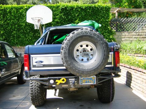 1985 Ford Bronco - The rear of a blue Ford Bronco is visible, a spare tire and a yellow ribbon sticker on the bumper.