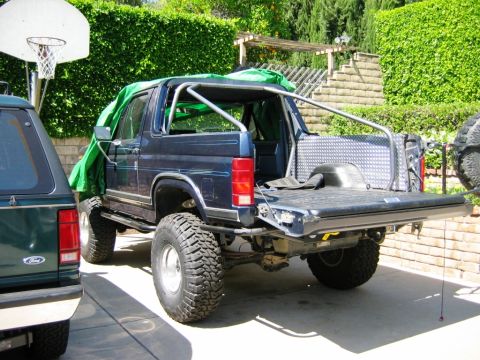 1985 Ford Bronco - Rear view of a modified 1985 Ford Bronco showing its roll cage and open tailgate, with another vehicle nearby.