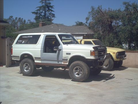 1990 Ford Bronco - Lifted white full-size Ford Bronco with a black "So-Cal Broncos" hood parked on a concrete driveway, driver's door open to show the gray cl…