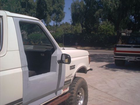 1990 Ford Bronco - Close view along the driver's side of a white full-size Ford Bronco with the door open and gray primer patches on the door and fender, park…