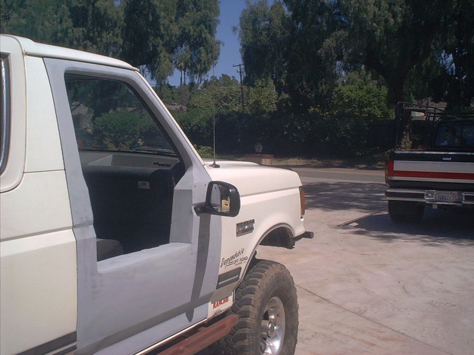 White Ford Bronco with primer patches and open door, mud tire, in a sunny driveway