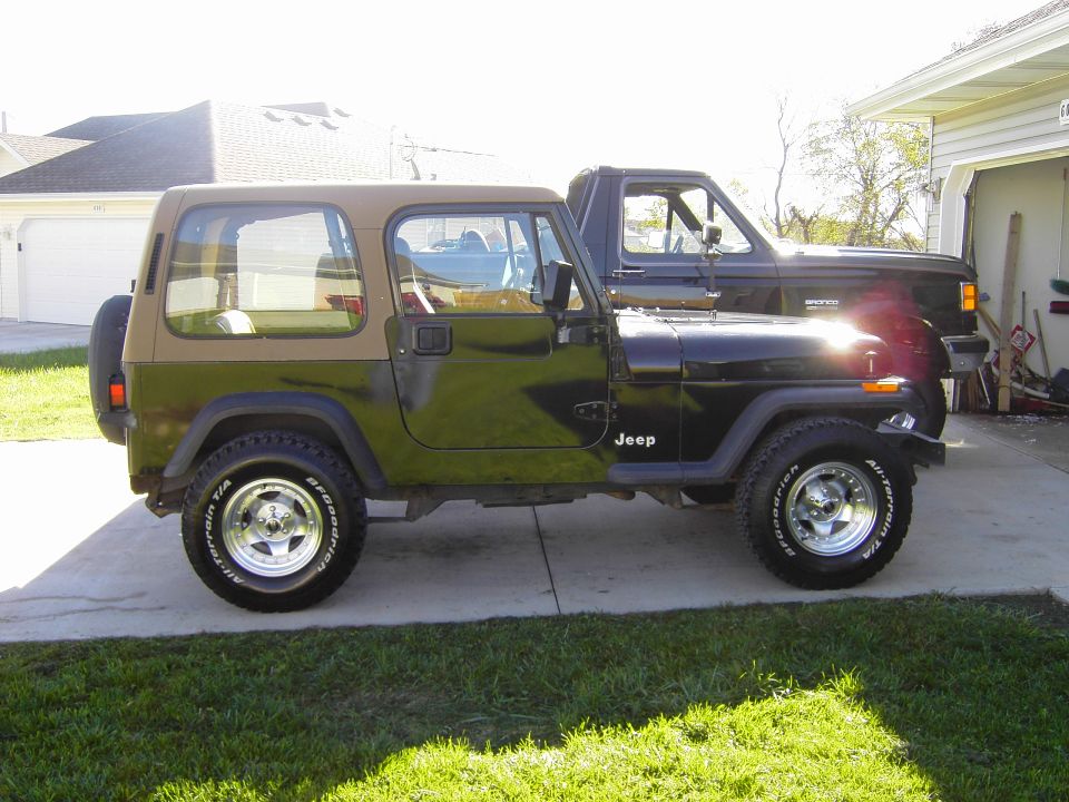 Two-door Jeep Wrangler with a tan hardtop and black lower body, parked next to a black pickup truck
