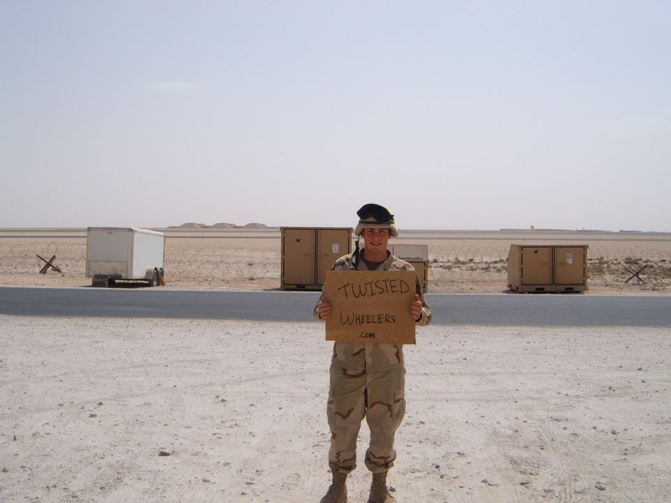 Soldier in desert uniform holds a cardboard sign that reads 'TWISTED WHEELERS.COM'