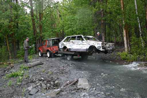 Red soft-top 4x4 towing a trailer with a stripped bullet-riddled car shell across a creek