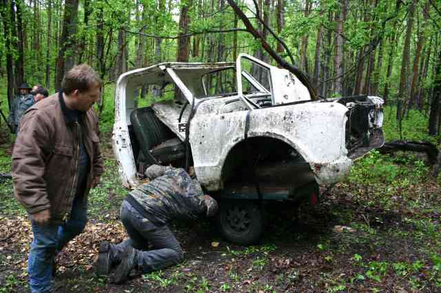 Stripped mud-covered white sedan shell in the woods with two men working on the rear