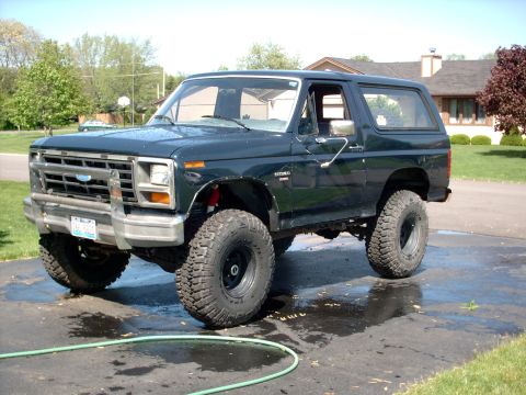 1986 Ford Bronco - A dark green/black 1980s Ford Bronco full-size SUV with a removable hardtop, lifted suspension and large off-road mud tires on black wheels…