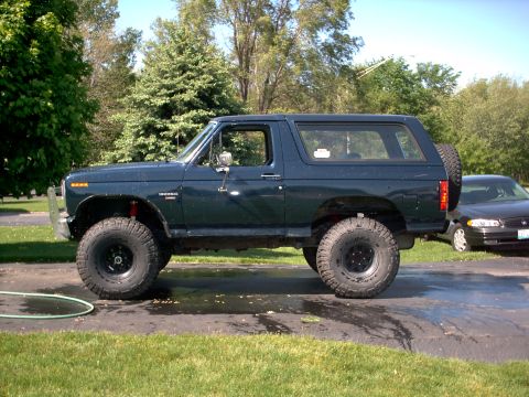 1986 Ford Bronco - Dark green/blue two-door Ford Bronco SUV shown in side profile, lifted with large off-road tires on black wheels and a rear-mounted spare t…