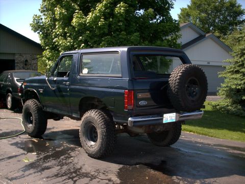 1986 Ford Bronco - Driver-side rear three-quarter view of a lifted dark navy blue full-size Ford Bronco on oversized mud-terrain tires with black wheels, a ch…