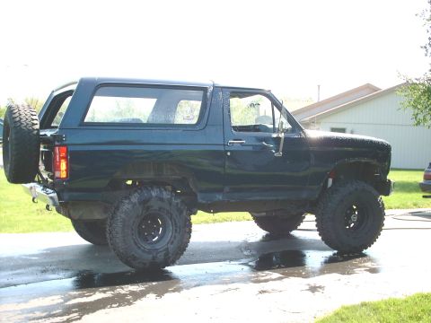 1986 Ford Bronco - A dark blue two-door 1986 Ford Bronco with a lift kit, oversized mud-terrain tires on black wheels, a rear tire carrier with spare, and a h…