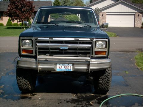 1986 Ford Bronco - Head-on front view of a freshly washed dark blue Ford Bronco sitting on mud-terrain tires in a wet suburban driveway. It has a chrome eggcr…
