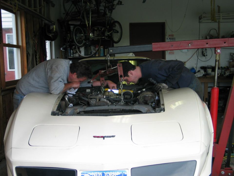 Two guys leaning over the engine bay of a white 1981 Chevrolet Corvette, working on it with an engine hoist overhead