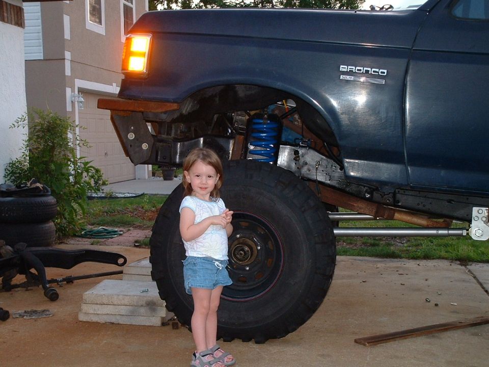 Dark blue Ford Bronco lifted on stands with exposed suspension, child standing by a large off-road tire.