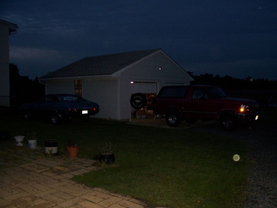 Dark muscle car and maroon lifted SUV parked by a white garage at dusk