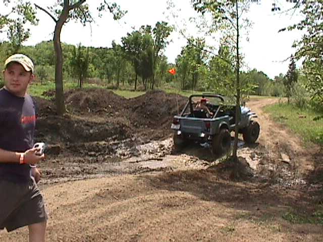 Guy in a baseball cap watches a Jeep Wrangler drive through mud on a dirt path