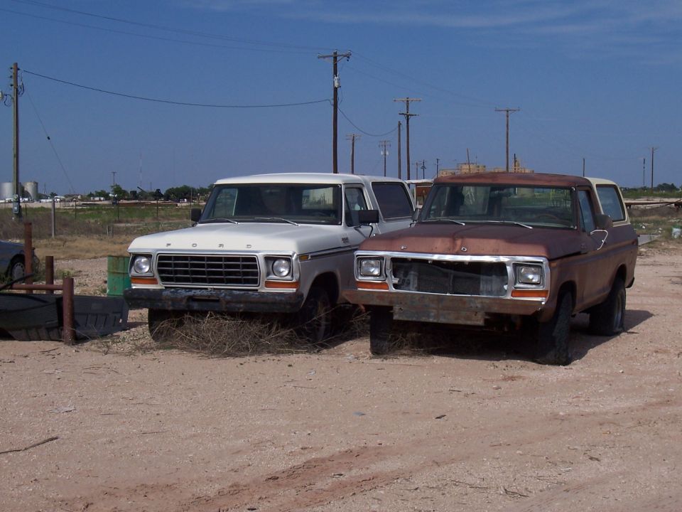 Two Ford Broncos parked in a lot, one white and one rusty brown, both with front damage