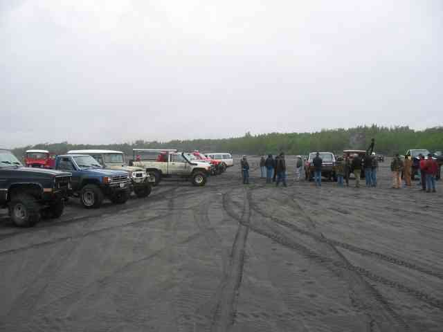 Row of lifted 4x4 trucks and SUVs parked on gray sand flats with people standing nearby