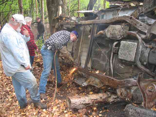 Wrecked vehicle chassis, heavily rusted and damaged, being cut with a torch by a person in a plaid shirt