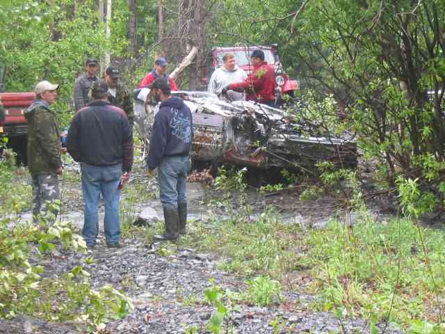 Several people stand around a heavily damaged vehicle in a forest clearing; another red vehicle is visible in the background