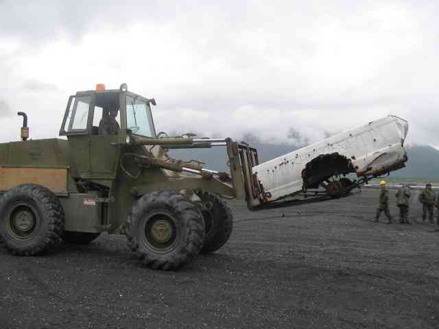 Forklift lifting a white vehicle body, with people in the background