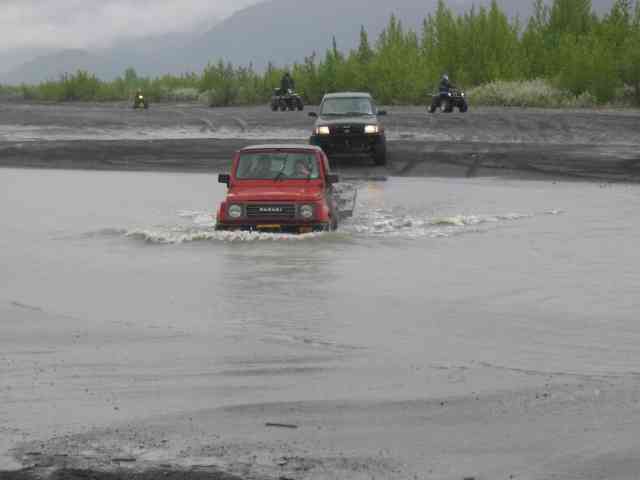 Red 1987 Suzuki Samurai driving through water with other vehicles in background