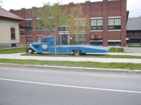 1979 Ford Bronco - A vintage blue tow truck with a long flatbed trailer is parked on the grass beside a brick building.