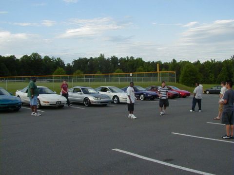 1994 Ford Probe - A group of people stand around several cars parked in an asphalt lot on a cloudy day.