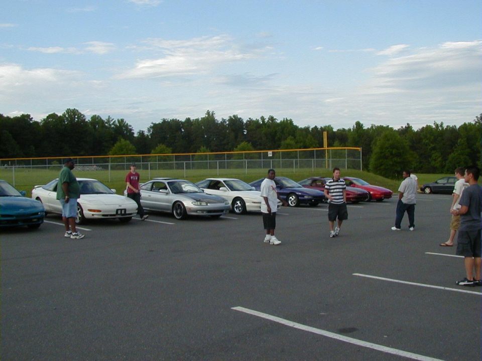 Several cars parked in a lot with people standing around. Some cars look like coupes