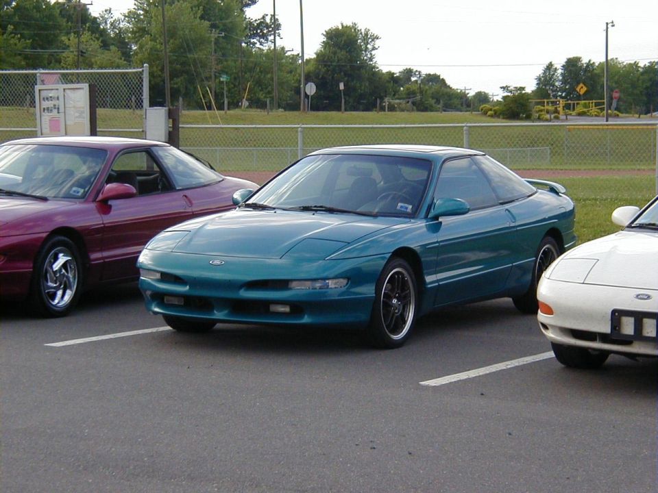 Teal Ford Probe parked in a lot, front 3/4 view. Another maroon Probe and white car are visible