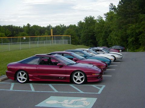 1994 Ford Probe - A line of Ford Probes, including a red GT model, are parked in a lot near a baseball field.