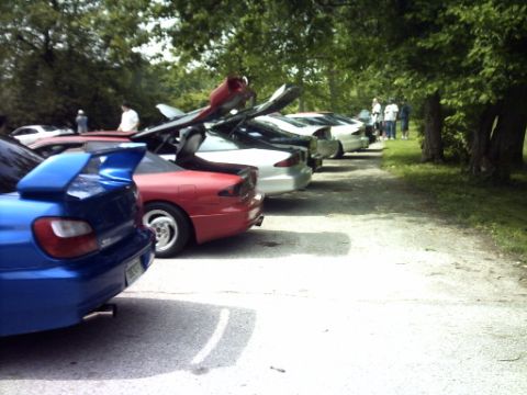 1995 Ford Probe - A gathering of cars, including a blue Subaru WRX, are parked in a row outdoors with trees in the background.