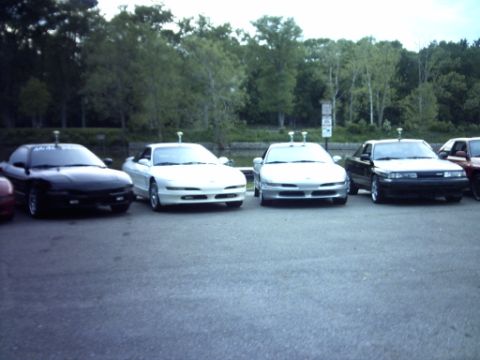 1995 Ford Probe - Multiple coupes are parked in a lot, with trees in the background and a water feature to the left.