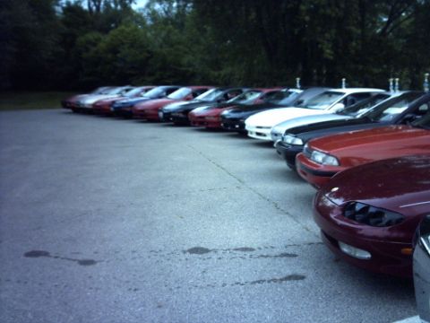 1995 Ford Probe - A line of cars is parked on an asphalt lot, with trees visible behind them.