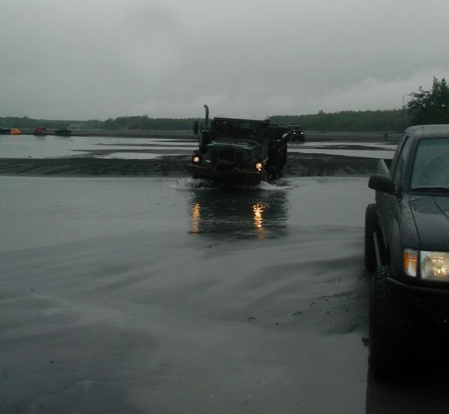Front view of a military truck driving through water, with a pickup truck on the right