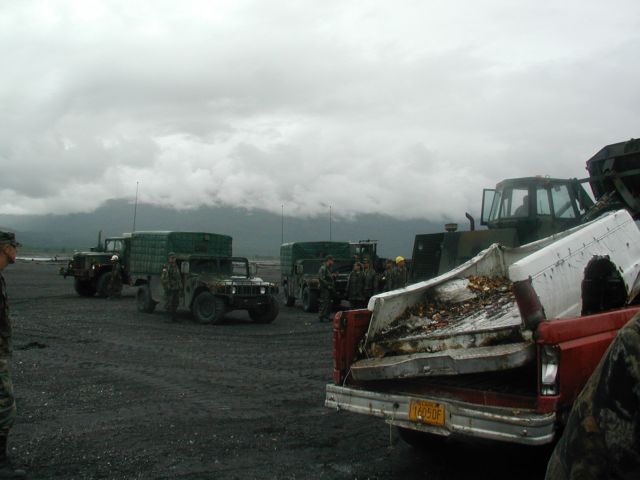 Military trucks and a bulldozer in a dusty landscape under a cloudy sky