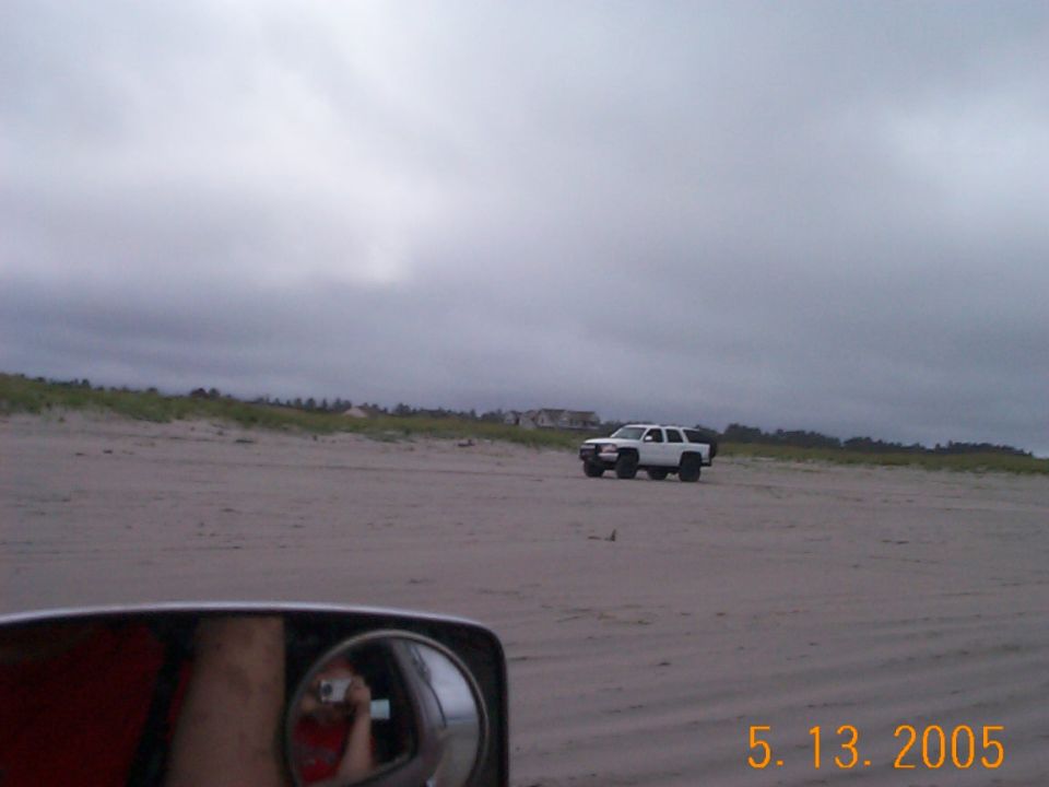 White SUV on a beach seen past a vehicle's side mirror under gray clouds