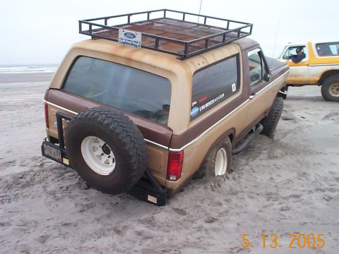 1983 Ford Bronco - A two-tone Ford Bronco is stuck in the sand on a beach, with another vehicle visible behind it.