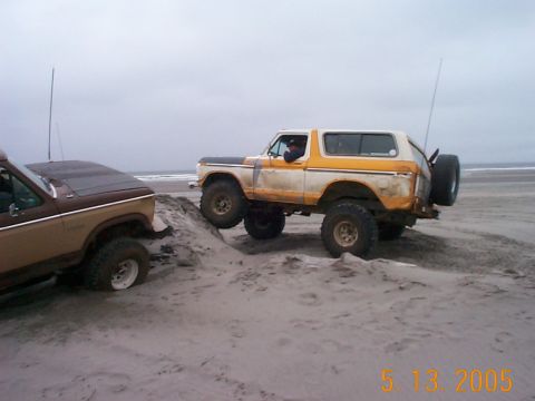 1983 Ford Bronco - Two off-road vehicles navigate a sandy dune near the ocean under an overcast sky.