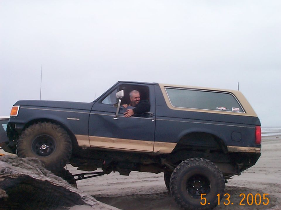 Man grinning from driver's seat of a lifted black-and-tan Bronco on a beach